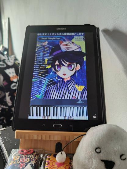 This is a photo taken of a tablet mounted on a wooden stand, which is positioned on a desk. The screen of the tablet is displaying the live broadcast of VTuber and musician Yamamii who is playing the piano for his Midnight Piano programme (something he does every Sunday). In front of the tablet I had positioned an onigiri plushie (made by Gumi Loki) and two GOATBED omamori. The background consists of a white wall and some things in the back, like a Halloween-themed blanket and a painting.