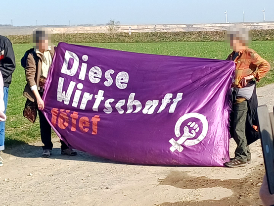Zwei Frauen* halten ein lola Banner: "Diese Wirtschaft tötet" mit feministischem Logo. Im Hintergrund der Tagebau Garzweiler II