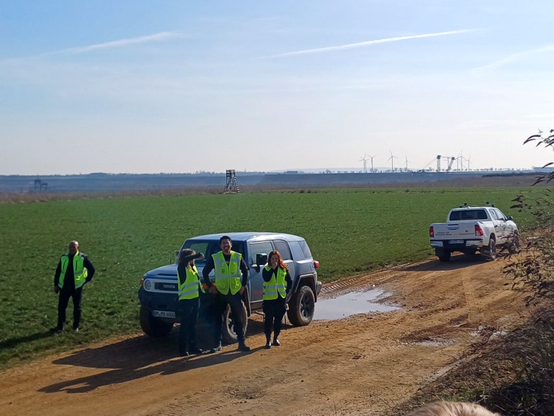 RWE-Secutirty mit zwei Werkschutz-Jeeps stehen mit gelben Warnwesten am Tagebaurand. Entfernt im Hintergrund - nahe dem ehemaligen Dorf Lützerath -  stehen ein Hochsitz, zwei Schaufelradbagger und mehrere Windräder.