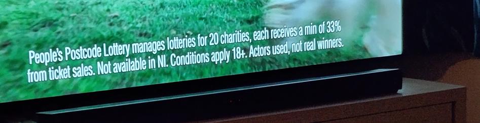 text at bottom of TV screen says
"People's Postcode lottery manages lotteries for 20 charities each receives a min of 33% from ticket sales not available in ni conditions apply 18 plus. Actors used, not real winners"