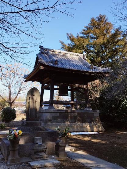 A Buddhist bell tower, a wooden gazebo with an ornate wooden roof, sits on a slightly elevated stone foundation. It's mostly in shadow so the tubular bell is only a contour against the sky. There are trees around it, some of them still bare, and to the left in front of it is a stone icon with flower offerings.