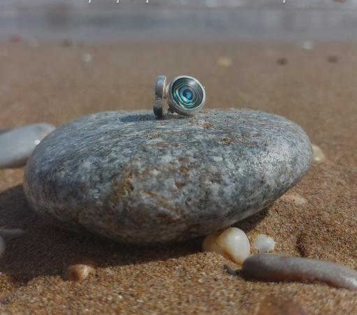a pair of round stud earrings. they are silver and has a round iridescent green stone. they are placed on a pebble in the beach