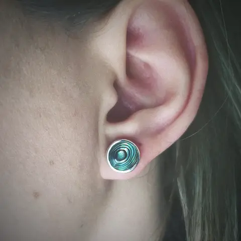 a round stud earring. it is silver and has a round iridescent green stone. it's worn by a woman