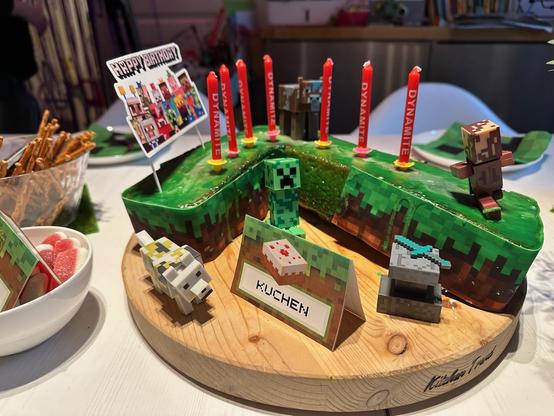 Ein Minecraft-Geburtstagskuchen mit roten "Dynamit"-Kerzen, Figuren- und Gegenstandsdekorationen, auf einem Holzbrett mit einem "Happy Birthday"-Schild und Snacks in der Nähe.
