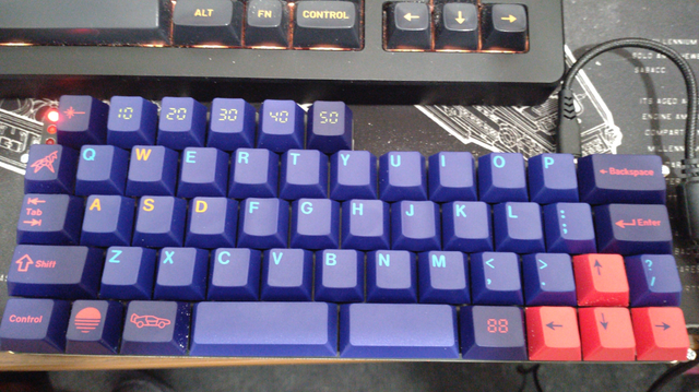 A 45% keyboard with four 12.75u rows and on top one 6u row slightly offset by three red LEDs on the left end.

It has cyan on a blue-ish purple alphas, red-ish pink on purple-ish dark blue modifiers and dark blue on red-ish pink arrow keys. The slash/question mark key is right of the curso up key.

There are several novelty keys used,.the left Alt key shows an icon of a DeLorean DMC12 timemachine from Back to the Future, the Windows key show a sunset icon, the tilde/backtick key a polygon unicorn icon, the Menu key show a 7-segment 88, and the Escape key shows a laser icon. The keys right of Escape in the top row are labeled 10 to 50 in a 7-segment style.