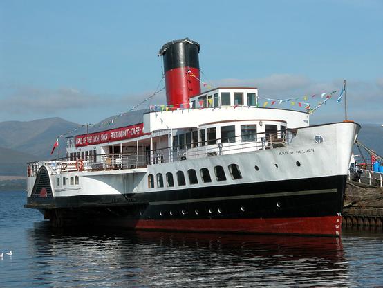 The paddle steamer Maid of the Loch. The image shows a front quarter view of the ship from its right side. It has a black and red lower hull and white upper hull and superstructure, with a red and black funnel. The ship is flying small flags fron strings running its length. The sky is blue.