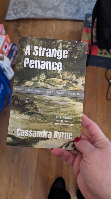 An author holds a proof for A Strange Penance. It feels exquisite to hold something physical like this in her hands. Cover art by Parris Ryan depicts a desert landscape, with an approach to a distant city.