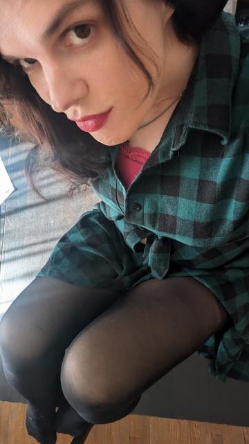 A transwoman poses for a thirst trap adjacent selfie. She is wearing black tights and a black and green checkered dress; she has long dark hair, brown eyes, and deliciously red lips. In this moment, she has temporarily vanquished her dysphoria.