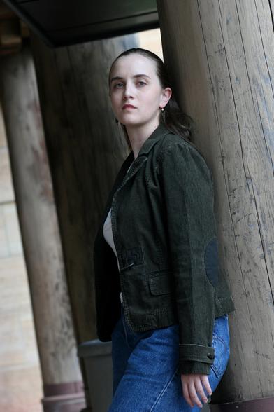 Photo of author Marie Brennan, a white woman with fair skin and long, dark hair pulled back. She wears blue jeans, a white shirt, a black denim fitted jacket, and small hanging earrings. She regards the camera with a dignified expression. Photo by Perry Reichanachter.