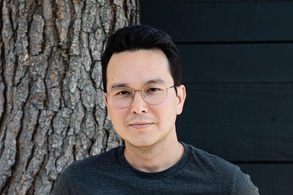 Writer Thomas Ha, a man of East Asian and white ancestry. He wears his almost-black hair short and swept off his forehead; he was a light mustache. He wears a charcoal-gray tee and gold-rimmed glasses. He regards the camera with an amused, somewhat skeptical expression.