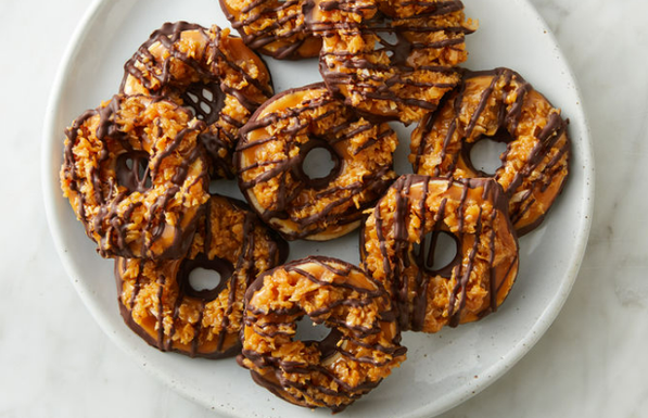 A white plate of Samoas Girl Scout cookies. Samoas are coconut based, have a hole in the middle, and are drizzled with chocolate.
