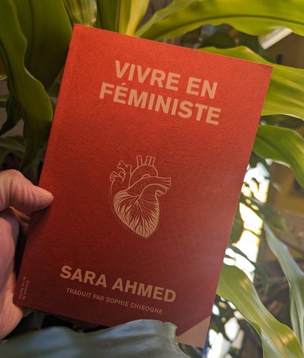 Le livre "Vivre en féministe" de Sara Ahmed dont la couverture est rouge texturé/métallique et affiche l'illustration d'un cœur biologique. Sur fond de plantes vertes.