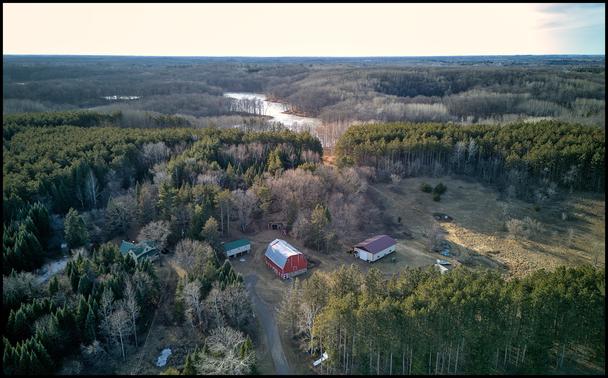 Aerial view of a red barn and associated house on the site of a small hill. The barn is surrounded by evergreen trees. The background is a bit hazy and we see a small lake/river winding through bare grey trees.