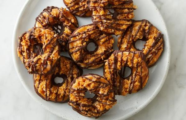 A white plate of Samoas Girl Scout Cookies. Samoas are brown, coconut-based, have a hole in the middle, and are drizzled with chocolate.