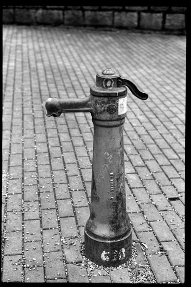 Black and white film photo. An old water pump on cobblestone place.