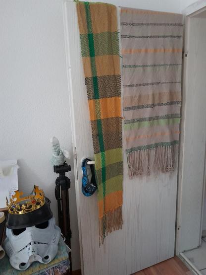 Deux tapis tissés main sèchent suspendus à une porte. L'un est très coloré, une alternance horizontale de bandes jaunes, vert pétrole, vert forêt, lime, khaki et beige foncé - moka, et une bande verticale vert forêt qui le traverse de part en part. Le tapis de gauche, plus large, a un fond de beige "nature" et est rayé des mêmes déclinaisons de verts, jaune et moka, mais en bandes beaucoup plus fines, et irrégulières. À gauche, devant la porte, sur une pile de cartons et d'écharpes, il y a un casque de storm trooper grandeur nature en attente d'un job de peinture, avec une couronne posée sur la tête.