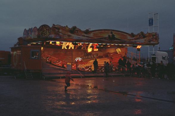 A child running in front of a ride under a rainy sky, lights reflecting on the ground