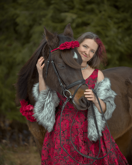 A photo of a woman and her horse. She wears a deep red dress covered with embroidered red floral patterns, and a fur shoulder drape with black-tipped grey fur. Her horse stands behind her, with his neck bent around her shoulder and head in front of her on one side. One of her hands is on his nose, and the other on his neck just behind the jaw. The horse has red flowers adorning his headband and chest strap.