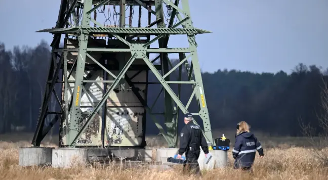 Zwei Polizist*innen vor der Basis eines großen 110-kV-Strommastes auf einem Feld. Der Mast hat auf ca. 4&nbsp;m Höhe Abwehrstacheln, die das Heraufklettern verhindern sollen. Unterhalb befindet sich eine Blechummantelung mit Brandspuren, hinter der die Kabel in den Boden verlaufen.

Foto: Sebastian Gollnow / dpa
