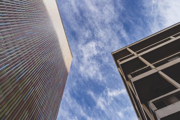 Looking up next to a building with coloured stripes to the left, and an apartment block to the right.
Blue sky with light cloud in the middle.