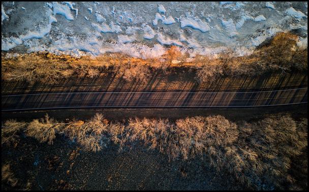 Looking straight down on a road between a frozen lake and a brown dry swamp. Small trees line the road on either side casting shadows across it.