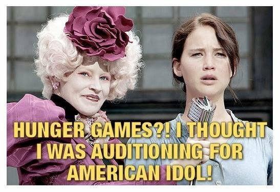 Two characters from the "Hunger Games" movie -- Effie Trinket in mauve, and Katniss Everdeen in light blue. They are on a stage, and Katniss is speaking into a microphone.
The text reads: "Hunger games?! I thought I was auditioning for American Idol!"