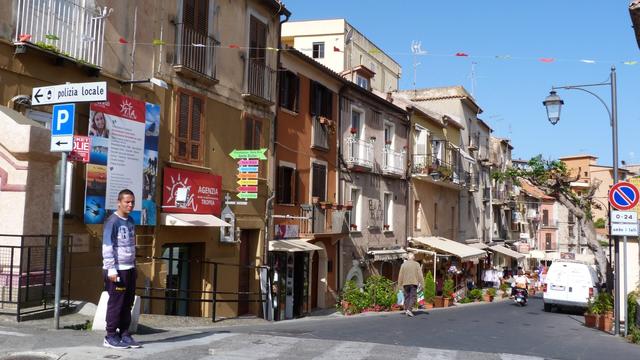 a sunlit street of houses in vernacular style, with painted fronts, slope down a gentle hill. a lone pedestrian waits at a crossing.