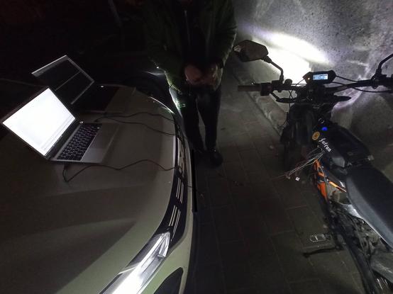 Bild außen bei Nacht.
Links ist ein Auto, dessen Scheinwerfer die Szene beleuchten, auf der Motorhaube stehen zwei Laptops.
Rechts ist ein E-Moped, aus dem Bereich des Akkus kommt ein Knäuel bunter Kabel. Ein paar Kabel gehen aus dem Knäuel vom Moped über diverse USB-Geräte zu den Laptops.