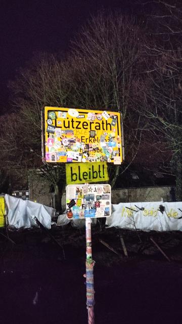 In nächtlicher Umgebung ein beleuchtetes Ortsschild "Lützerath - Stadt Erkelenz" mit vielen wilden Aufklebern, darunter ein ebenfalls gelbes Schild mit schwarzer handgeschriebener Aufschrift "bleibt". Aufgenommen am 10.01.2023. Kurz danach wurde Lützerath geräumt.