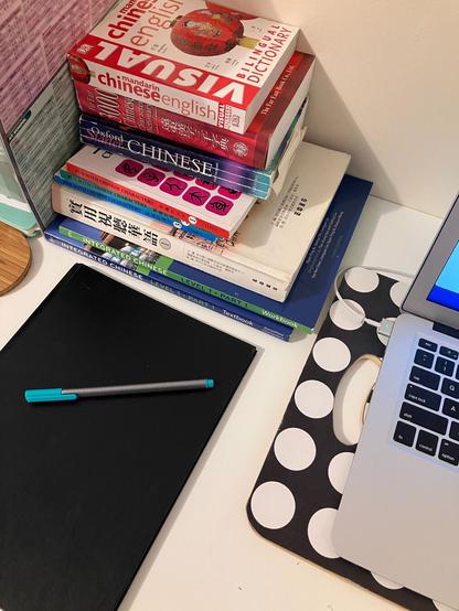 A desktop with a notebook and pen, a laptop, and a stack of Chinese dictionaries and textbooks.