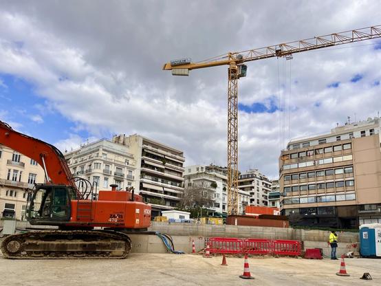 A crane looms over an apartment building, the arm parallel with the roof. On the opposite side of the frame, a machine with treads an an extended arm. It’s the construction site of the new metro station in kolonaki square, Dort and concrete wall and portable toilets all surrounded by tall buildings.
