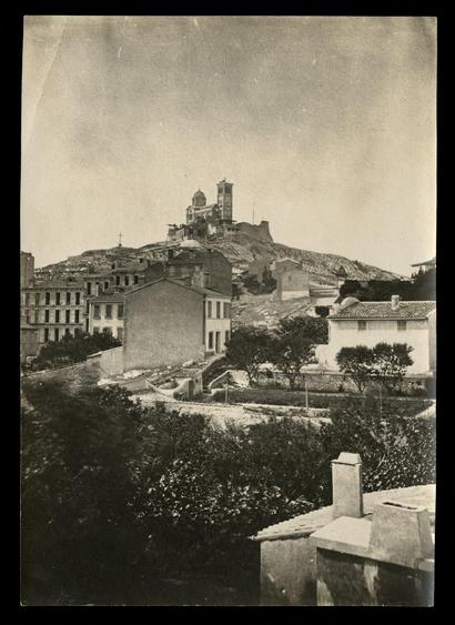 Photographie en noir et blanc montrant Notre-Dame de la Garde de Marseille encore en construction. La statue de la vierge est absente. Au premier plan du bâti avec quelques maisons et immeubles.