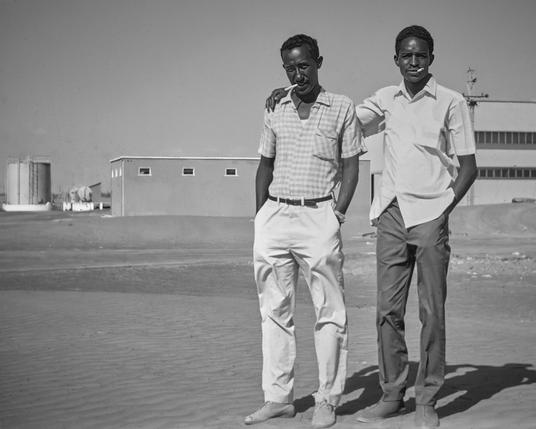 This is a black and white photograph of two men from 1969. They are standing side by side on a sandy ground with a large industrial building and a power plant in the background. The man on the left is leaning slightly towards his friend, relaxed, with a soft smile on his face. He is dressed in a light-colored, checked, short-sleeved shirt and white trousers. The man on the right stands tall and straight, his expression serious, and he wears a crisp white shirt with dark trousers. Both are wearing shoes suited for outdoor work. The clear sky above and the buildings behind them tell a story of industrial work in the vast desert of Al Ain, UAE. The photograph speaks to the era's style and the country's development phase, captured by the enduring qualities of Kodachrome film and a Soviet-era Cosmic 35 camera