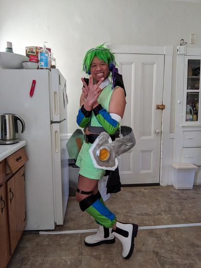 Me cosplaying Reina Prowler's Strahl outfit from Macross Delta: Green spiky wig, green vest and shorts, blue and green striped arm and leg warmers, and giant headphones around my waist.