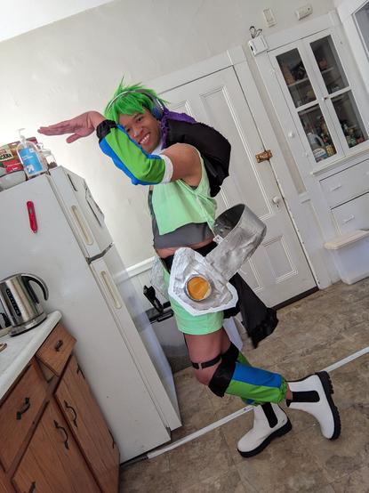Me cosplaying Reina Prowler's Strahl outfit from Macross Delta: Green spiky wig, green vest and shorts, blue and green striped arm and leg warmers, and giant headphones around my waist.