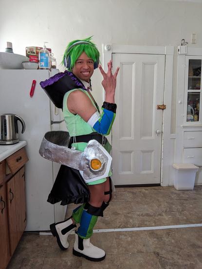 Me cosplaying Reina Prowler's Strahl outfit from Macross Delta: Green spiky wig, green vest and shorts, blue and green striped arm and leg warmers, and giant headphones around my waist.
