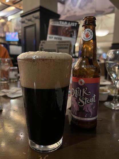 A bottle of Lefthand Nitro Milk Stout with a goofy new purple and white label. A well poured pint glass with a tall head stands to the left.