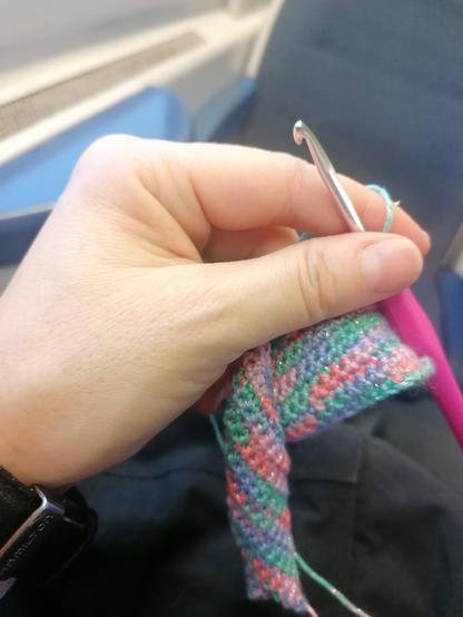 La mia mano sinistra regge il lavoro iniziato all'uncinetto: il filato è rosa, aragosta, verde, glicine glitterato; l'uncinetto ha l'impugnatura rosa ciclamino. Nell'immagine ci sono anche parte del sedile di fronte a me, vuoto, e del finestrino del treno su cui sto viaggiando