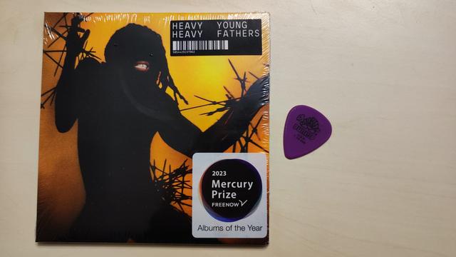Eine CD Hülle, noch in Folie, mit zwei Aufklebern darauf. Das Cover zeigt den Schattenriss einer tanzenden Person, die Arme leicht erhoben, der Mund ist hervorgehoben, zeigt hell die Zähne, der Hintergrund ist Orange und weist schwarze Linien auf. Auf dem oberen Aufkleber steht "Heavy Heavy, Young Fathers", darunter ein Barcode. Auf dem unteren Aufkleber, in einem runden Kreis "2023 Mercury Prize".
Neben der CD liegt ein violettes Gitarren Pick.