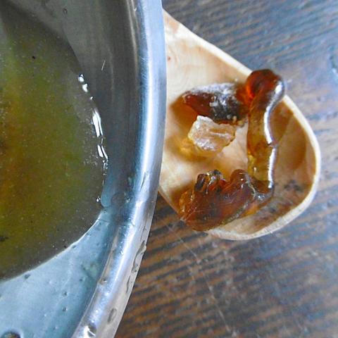 In einer Schüssel ist gelöstes Kirschgummi zu sehen. Es sieht gelblich dick aus, ähnlich wie flüssiger Honig. Neben der Schüssel liegen auf einem Holzlöffel noch einige Stücke Kirschgummi.