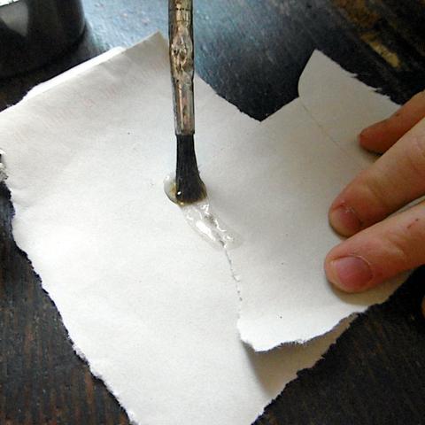 Eine Hand hält zwei Stück Papier fest. Ein Pinsel verstreicht flüssigen Gummikleber auf und zwischen die Papiere.