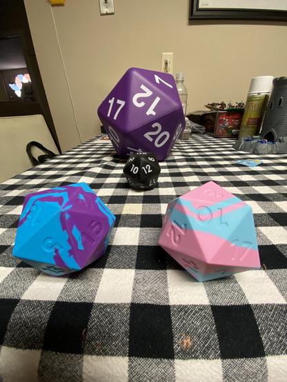 Three oversized polyhedral dice on a checkered tablecloth with a blurred background suggesting a room interior.