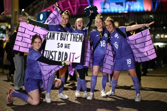 Eine Gruppe von Fans steht vor einem Stadion und hält ein Stoffbanner hoch auf dem steht: "We can't buy it, so we just did it" mit dem Nike Haken dahinter. Sie tragen selbstgemachte violette Trikots von Australiens Nationaltorhüterin Mackenzie Arnold und halten außerdem weitere Stoffbanner mit einem Mauerwerkmuster darauf.