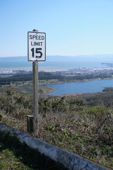 The image shows a vertical white speed limit sign with black lettering mounted on a wooden post. The sign indicates a speed limit of 15. The background behind the sign shows the calm water of a lake, with rolling hills and trees visible in the distance. (From Gemini.)