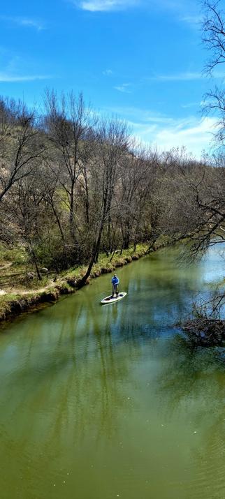 A man in a light blue shirt, gray pants tucked into black boots, on a standup paddleboard in a river with green water, the river meanders in a forest by a hill.