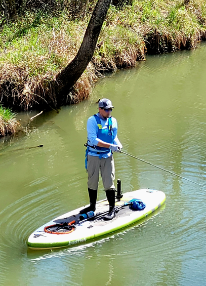 A man in a light blue shirt, gray pants tucked into black boots, fishing while on a standup paddleboard in a river with green water.
