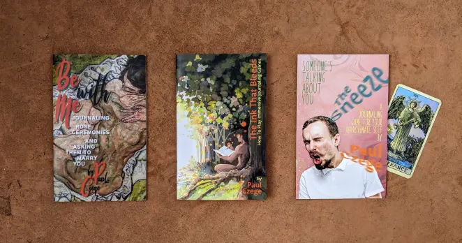 Copies of my zines, The Ink That Bleeds, about how to play immersive journaling games, Be With Me, a journaling game about your time on a reality dating show hoping to find love, and The Sneeze, a journaling game about a character you know from another game talking about you when you're not there.