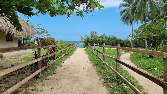 A fenced trail leads into the sea and the blue sky, surrounded by vegetation. A thatched hut stands on the left.
