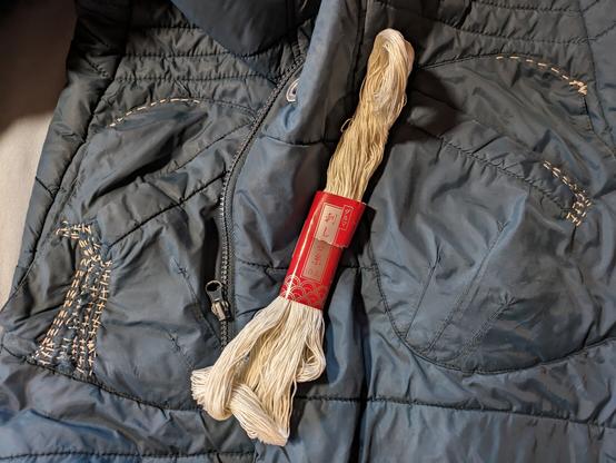 Die Jacke in Draufsicht. Auf beiden Seiten sind die Jackentaschen sichtbar mit weissem sashikogarn bestickt und geflickt. Das naturweisse Garn liegt als Strange mit japanischer rotgoldener Banderole in der Mitte.