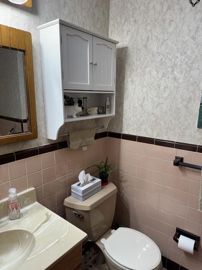 A bathroom interior with beige and brown tiles, featuring a white cabinet, mirror, sink, toilet, and various toiletries.